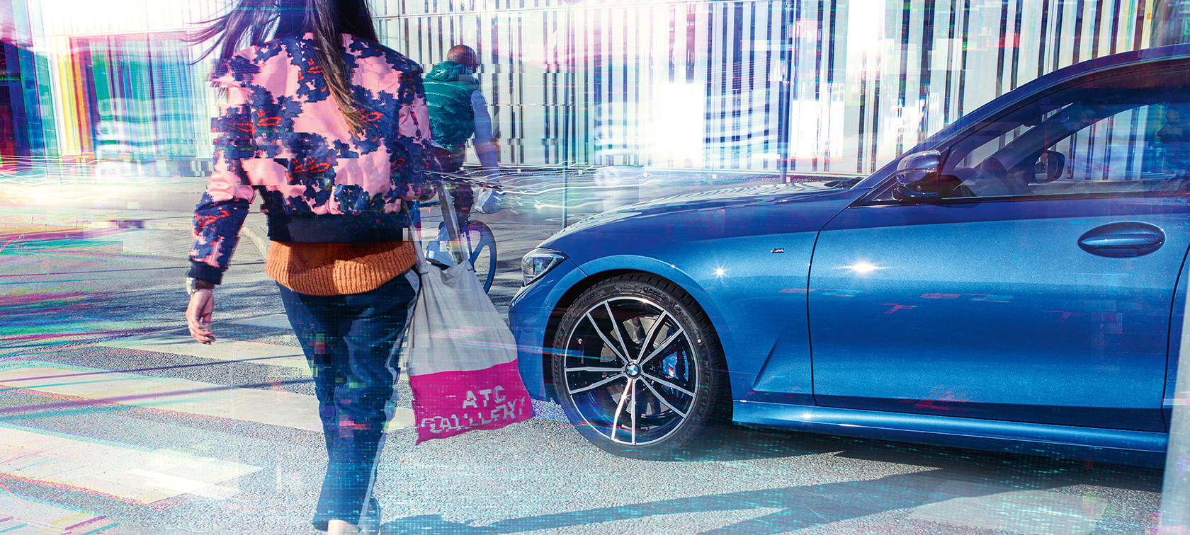 BMW Série 3 Berline, vue latérale de trois-quarts. Vue latérale de trois-quarts de la BMW Série 3 Berline sur l’asphalte devant un décor urbain avec passants.